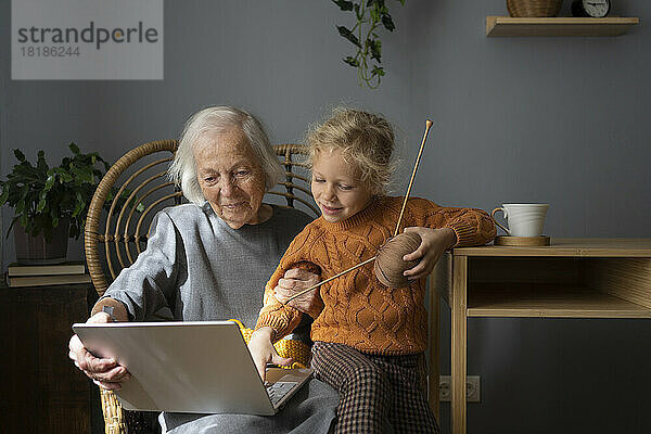 Lächelnde Großmutter und Enkelin lernen zu Hause das Stricken per Laptop