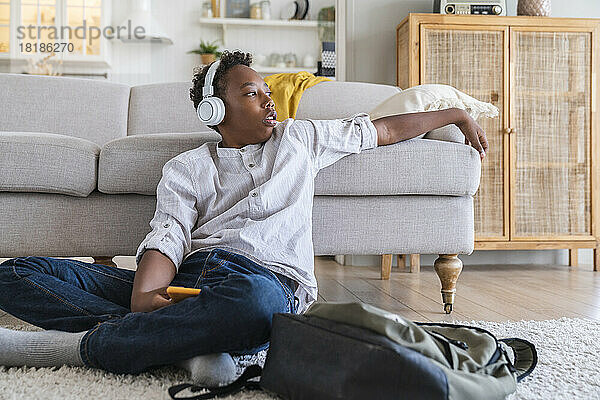 Nachdenklicher Junge mit Kopfhörern  der zu Hause auf dem Teppich Musik hört