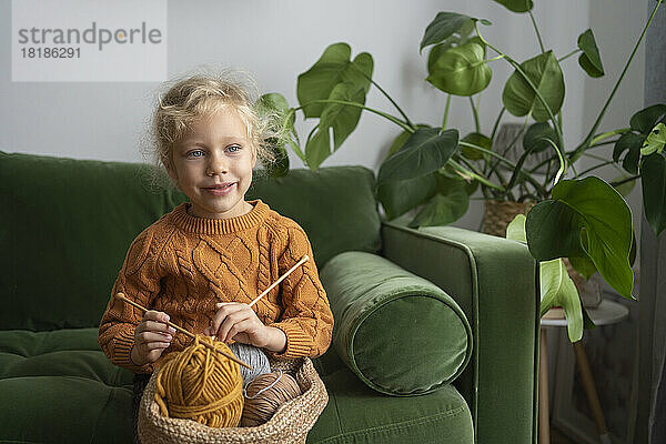 Mädchen hält einen Korb mit Wollknäueln und Stricknadeln auf dem heimischen Sofa