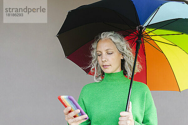 Frau mit buntem Regenschirm benutzt Smartphone vor der Wand