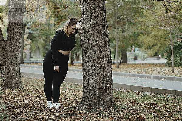 Erschöpfte Frau lehnt im Herbstpark an Baumstamm