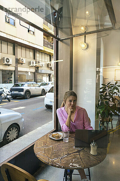 Lächelnder junger Freiberufler sitzt mit Laptop am Tisch im Café