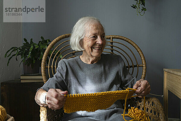 Glückliche ältere Frau mit Strickwolle und Nadel auf dem Stuhl zu Hause