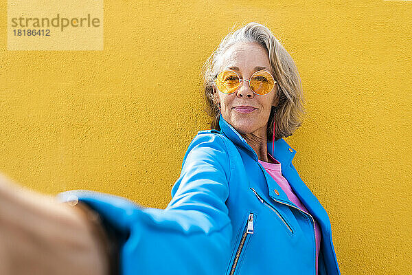 Smiling mature woman taking selfie in front of yellow wall
