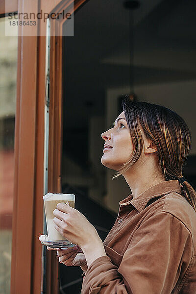 Nachdenkliche junge Frau mit einem Glas Cappuccino steht in der Tür eines Cafés