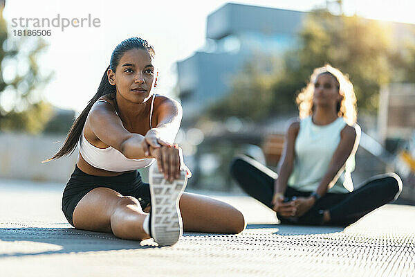 Teenage girl practicing stretching exercise with friend sitting in background on sunny day