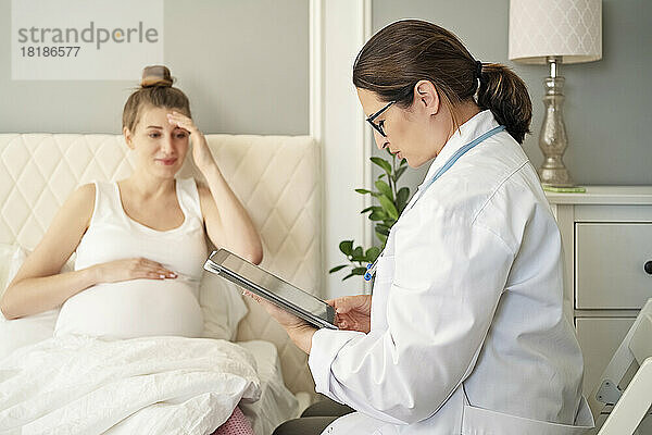 Arzt mit Tablet-PC berät schwangere Frau zu Hause im Bett