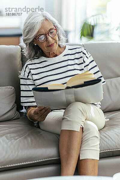Frau liest ein Buch und sitzt mit gekreuzten Beinen zu Hause auf dem Sofa