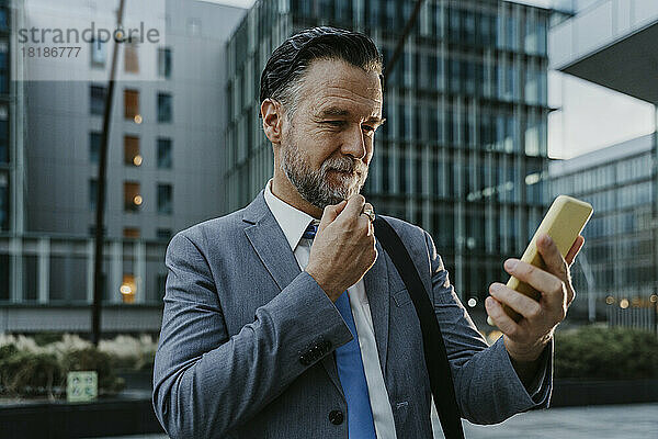 Nachdenklicher reifer Geschäftsmann blickt vor dem Gebäude auf sein Smartphone