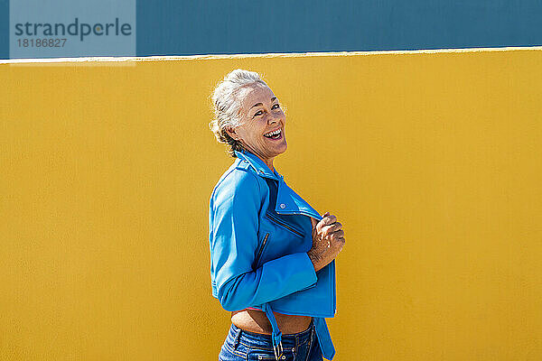 Glückliche Frau in blauer Lederjacke vor farbiger Wand