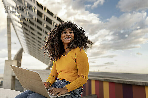 Glückliche junge Frau sitzt mit Laptop unter bewölktem Himmel