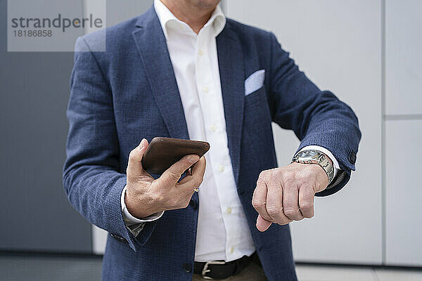 Geschäftsmann hält Mobiltelefon in der Hand und überprüft die Uhrzeit auf der Armbanduhr vor grauer Wand