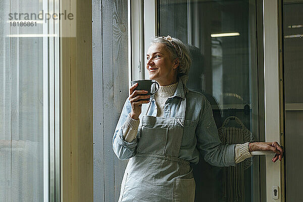 Lächelnde Handwerkerin hält Kaffeetasse am Eingang in der Werkstatt