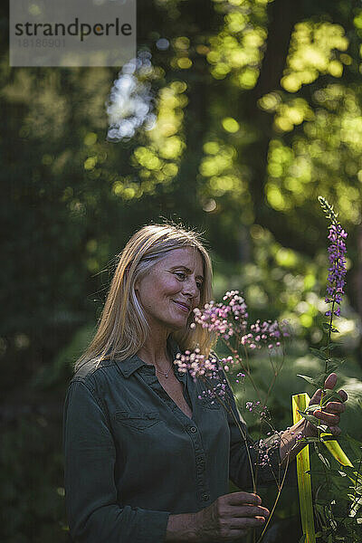 Lächelnde reife Frau berührt blühende Pflanzen im Garten