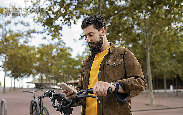 Junger Mann benutzt Smartphone und steht mit Fahrrad vor Baum