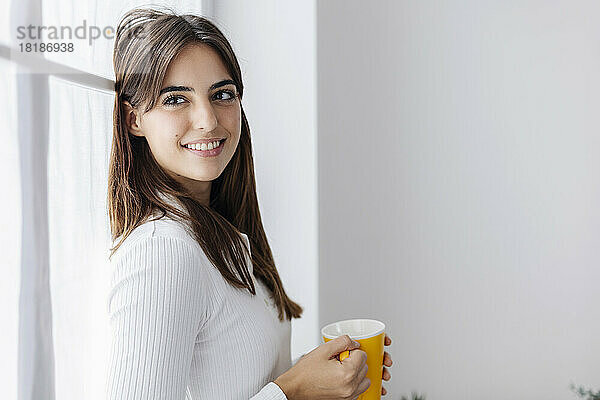 Lächelnde Frau mit Kaffeetasse lehnt am Fenster