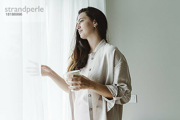 Nachdenkliche Frau mit Kaffeetasse und Blick durch das Fenster zu Hause