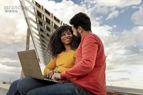 Glückliches junges Paar sitzt mit Laptop unter bewölktem Himmel
