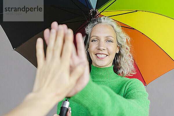 Lächelnde Frau mit buntem Regenschirm  die einem Freund ein High-Five gibt