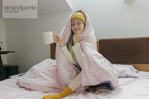 Junge Frau hält eine in eine Decke gewickelte Kaffeetasse zu Hause im Bett