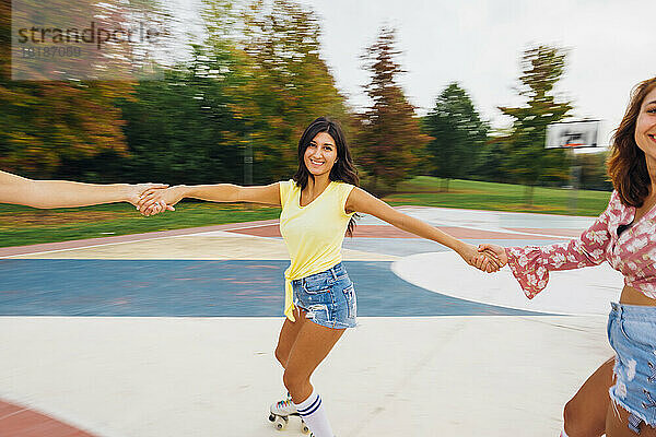 Lächelnde Frau hält Händchen von Freunden beim Rollschuhlaufen auf dem Sportplatz