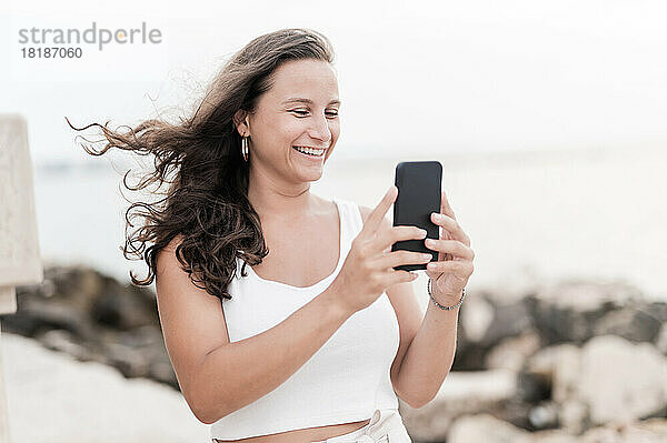 Lächelnde Frau fotografiert am Wochenende mit ihrem Smartphone am Strand