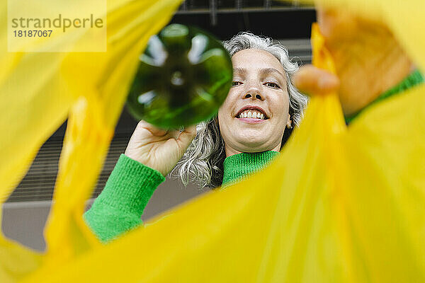 Lächelnde Frau steckt grüne Flasche in gelbe Plastiktüte