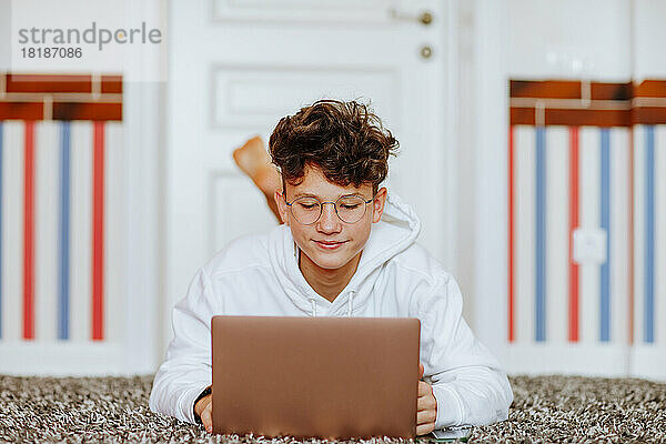 Lächelnder Junge liegt zu Hause auf dem Teppich vor dem Laptop