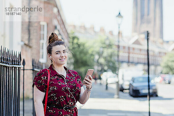 Porträt einer Frau  die mit Kopfhörern und Mobiltelefon auf dem Bürgersteig steht  Liverpool  Großbritannien