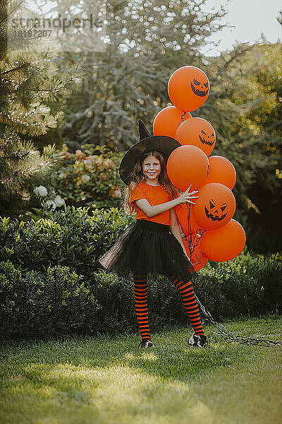 Lächelndes Mädchen mit Hexenhut und orangefarbenen Luftballons im Hinterhof