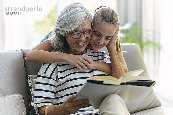 Lächelndes Mädchen umarmt Großmutter und liest zu Hause auf dem Sofa ein Buch