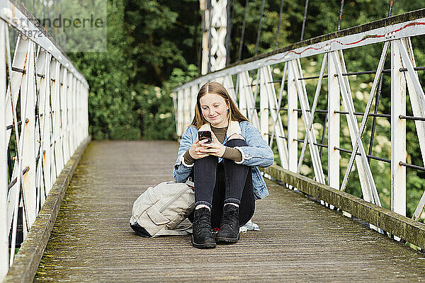 Teenager-Mädchen sitzt auf einer Brücke und benutzt Smartphone