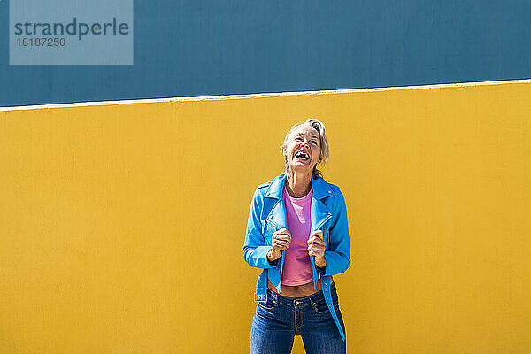 Glückliche reife Frau in blauer Lederjacke vor farbiger Wand