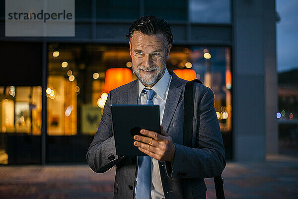 Lächelnder reifer Geschäftsmann steht mit Tablet-PC auf dem Fußweg