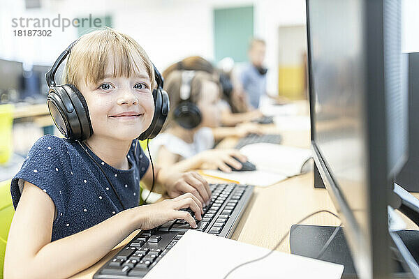 Lächelndes Mädchen mit Pony und kabelgebundenen Kopfhörern sitzt im Computerunterricht in der Schule