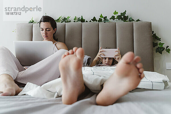 Frau mit Laptop sitzt neben Mädchen und benutzt Smartphone  das zu Hause im Bett liegt