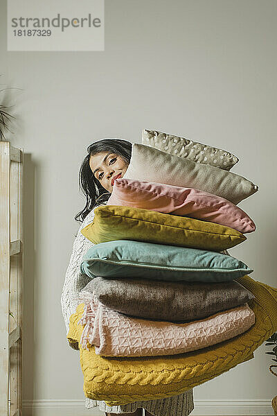 Smiling woman holding pillows and blankets in front of wall at home
