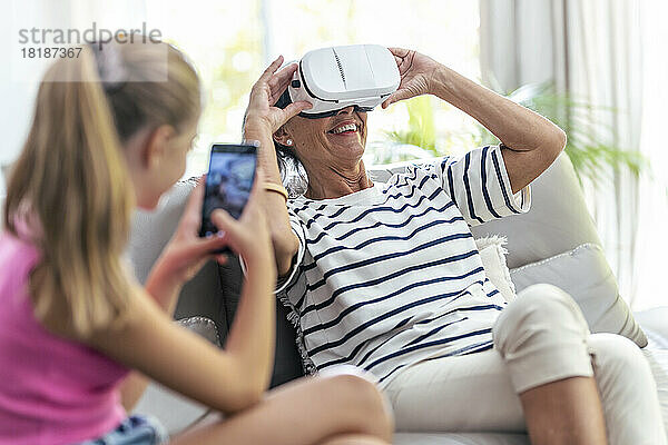 Enkelin fotografiert ältere Frau  die zu Hause ein Virtual-Reality-Headset genießt