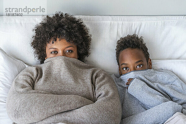 Mutter und Sohn bedecken den Mund mit Pullover und liegen zu Hause im Bett