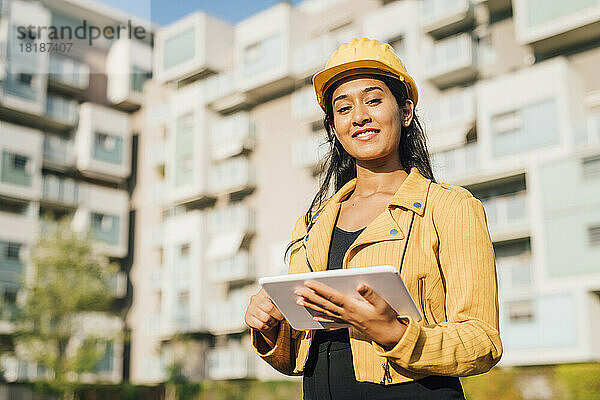 Glückliche Frau mit Tablet-PC und Bauarbeiterhelm  die an einem sonnigen Tag steht