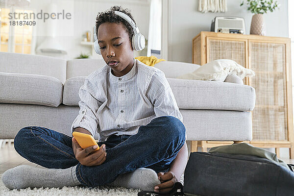 Junge trägt Kopfhörer und hört Musik  während er zu Hause mit dem Mobiltelefon sitzt