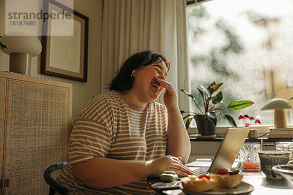 Fröhliche junge Frau  die den Mund mit den Händen bedeckt  während sie am Tisch am Fenster sitzt