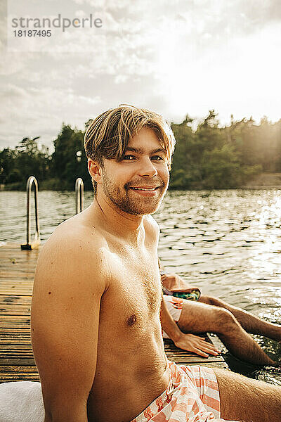 Lächelnder Mann ohne Hemd  der im Urlaub auf dem Bootssteg sitzt