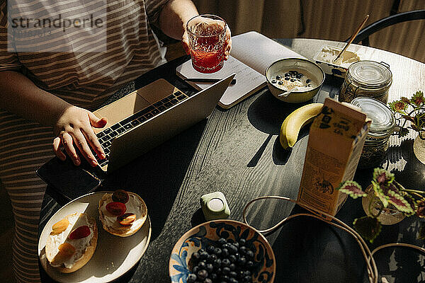 Hohe Winkel Ansicht der Frau hält Trinkglas bei der Verwendung von Laptop mit Frühstück auf dem Tisch zu Hause
