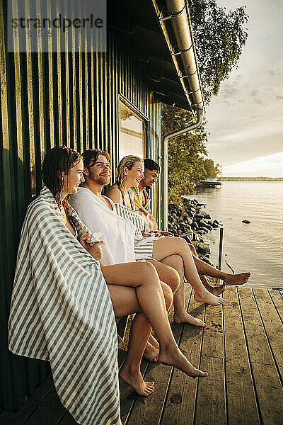 Männliche und weibliche Freunde sitzen bei Sonnenuntergang in der Nähe einer Hütte mit übereinander geschlagenen Beinen