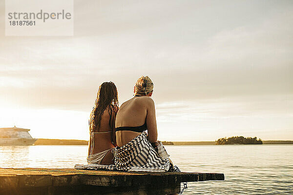 Rückansicht von Freundinnen  die bei Sonnenuntergang auf einem Steg sitzen und auf den See blicken