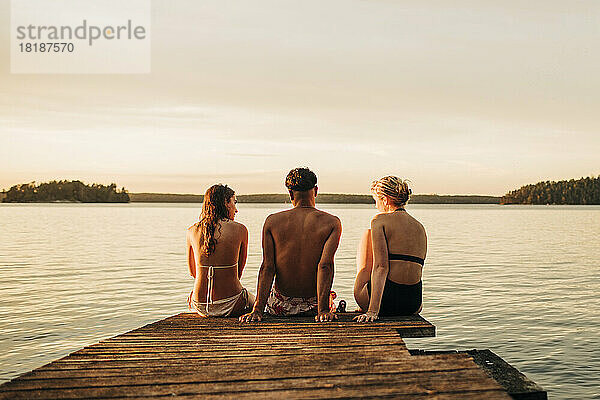 Rückansicht von männlichen und weiblichen Freunden  die im Urlaub auf einem Steg am See sitzen