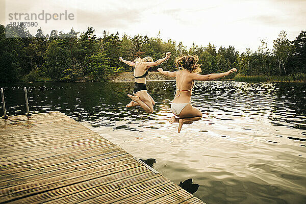 Freundinnen im Bikini  die im Urlaub mit ausgestreckten Armen in den See springen