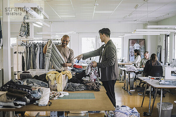 Seitenansicht eines männlichen Modedesigners  der auf einen Kollegen zeigt  der in einer Werkstatt einen Pullover hält