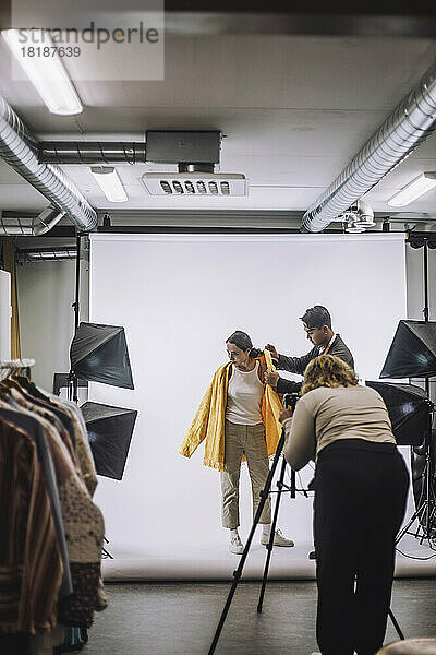 Fotograf fotografiert Designer hilft Modell trägt Jacke vor weißem Hintergrund im Studio